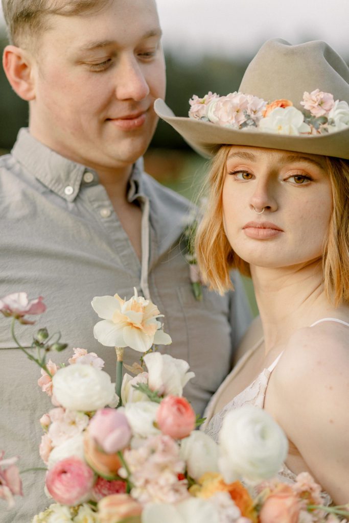 Bride in cowboy hat with spring florals looks at the camera, while her husband looks down at her lovingly.