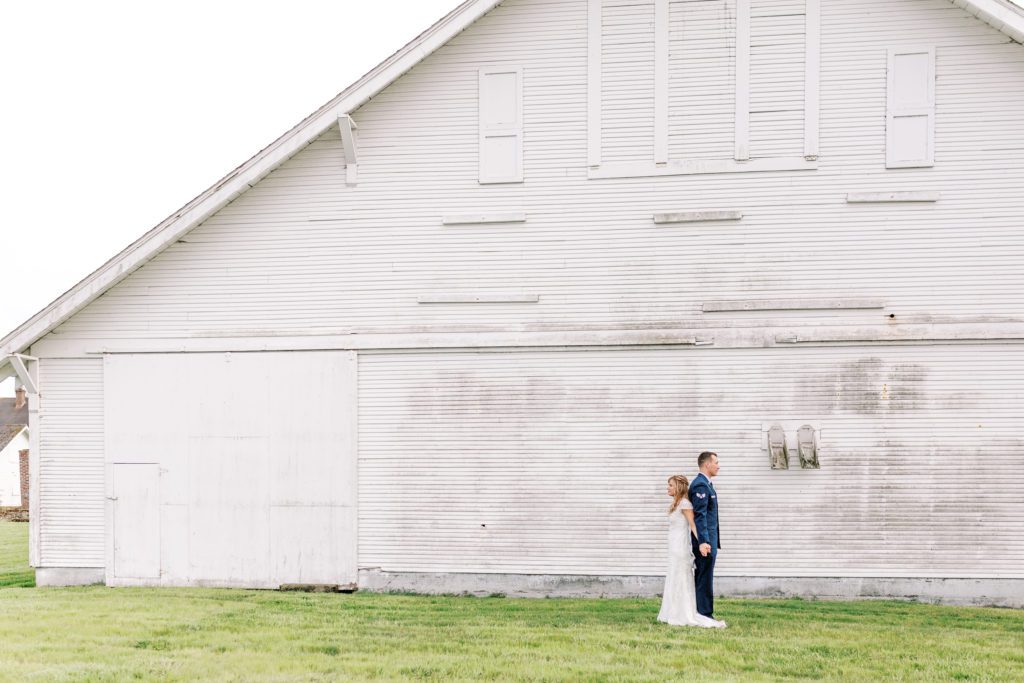 newlyweds stand in front of a white barn with fresh green grass in washington