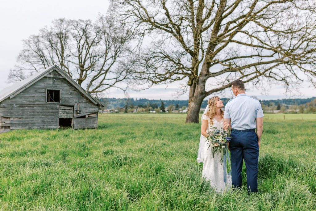 bride and groom embrace in a field of green grass and an old barn